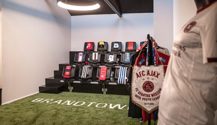 Showroom med gräsmatta och en liten läktare. Halsdukar med olika fotbollslag ligger på läktarplatserna och ställ med kläder från Brandtowns kunder hänger utmed väggarna.