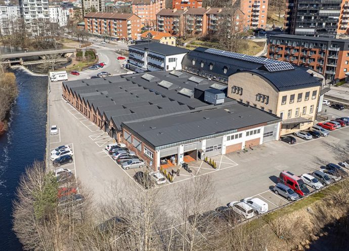 Drönarbild över Forsen sett från baksidan med stora portar för verkstadslokaler. Ån Viskan syns flyta förbi i två väderstreck.